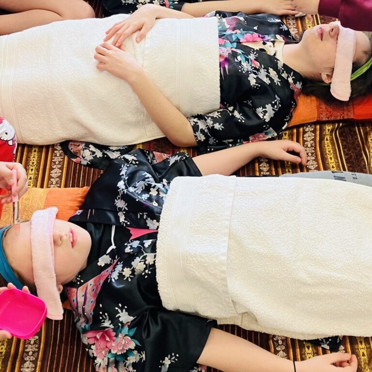 Two girls lying down with robes and an eye mask on, one is having a face mask applied to her face.