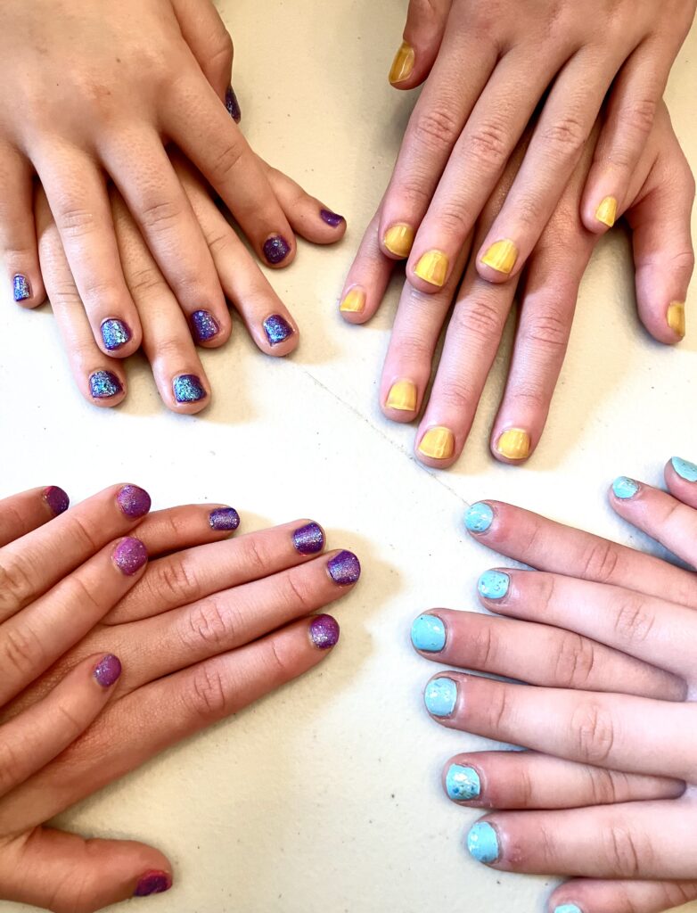 Four pair of hands displying their nail polish. They have one hand on top of the other. There is yellow, purple and light and dark blue nails.