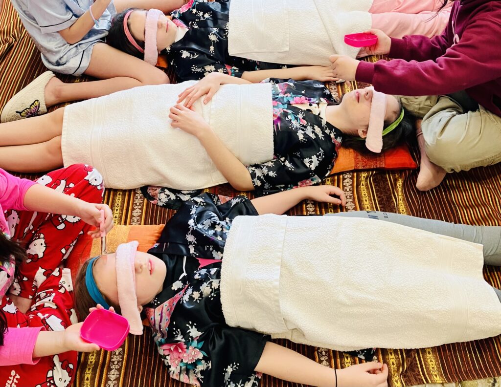 Three girls lying down top to tail having face mask applied. They are all wearing gowns with white towels over them and pink eye masks.