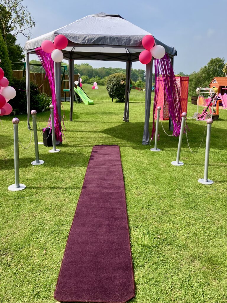 Maroon runner laid onto of a grass lawn with a silver gazebo in the background. It's a bright sunny day.